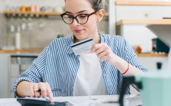 A young woman holding a credit card and using a calculator while sitting at a kitchen table