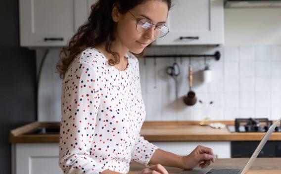 Female standing in home kitchen doing accounting work from home, use calculator fill data results on laptop application