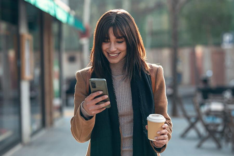 Woman walking on a sidewalk, looking at her phone, holding a coffee cup