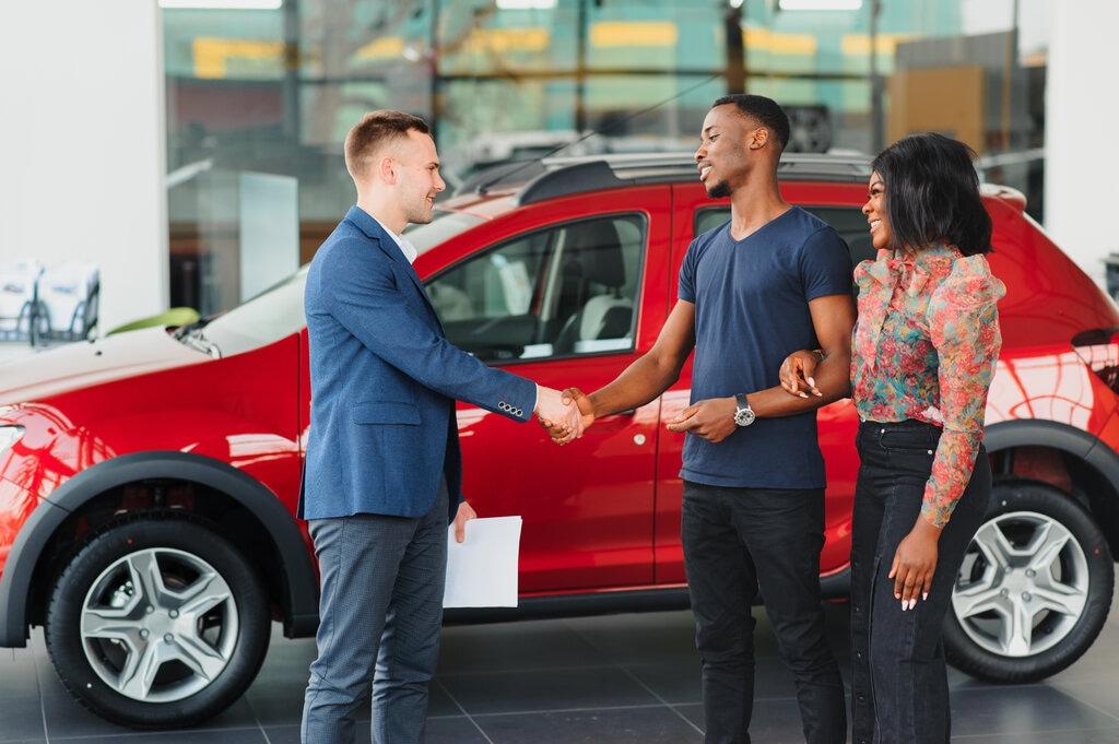 Couple standing in front of red car shaking salesman's hand