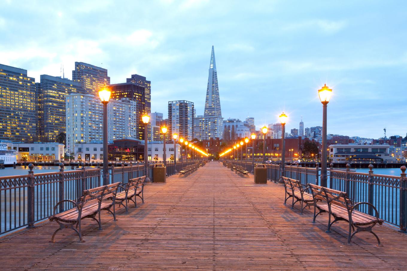 A wooden pier lined with glowing streetlamps leads toward the San Francisco skyline at dusk, with the Transamerica Pyramid rising in the background.