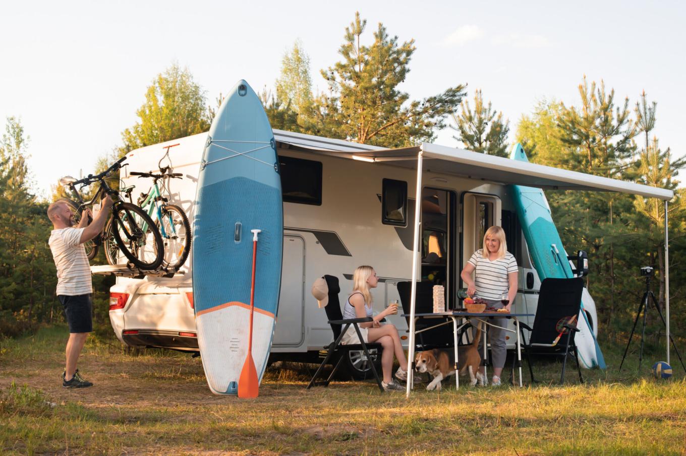 A family relaxes beside a camper van at a forest campsite, with bicycles, paddleboards, and outdoor gear set up for a day of adventure.
