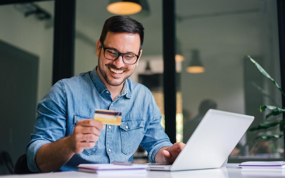 Happy smiling young adult man looking at his credit card while online shopping