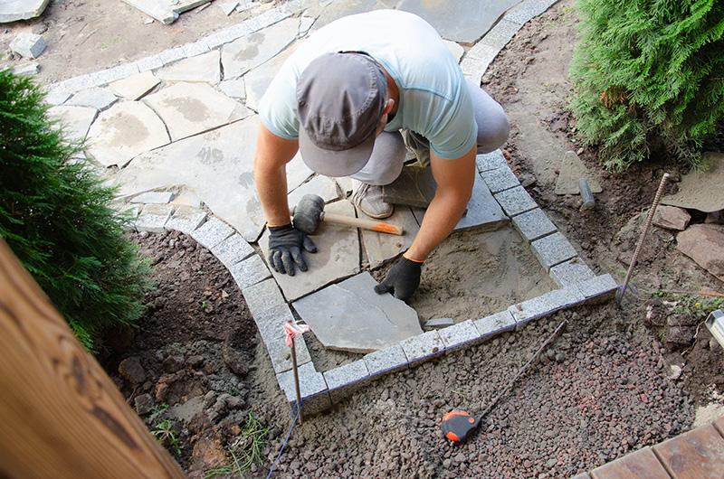 A person places a tile along a garden path with tools and plants nearby.