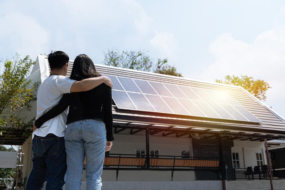 A couple stands with their arms around each other, looking up at solar panels installed on the roof of their home.
