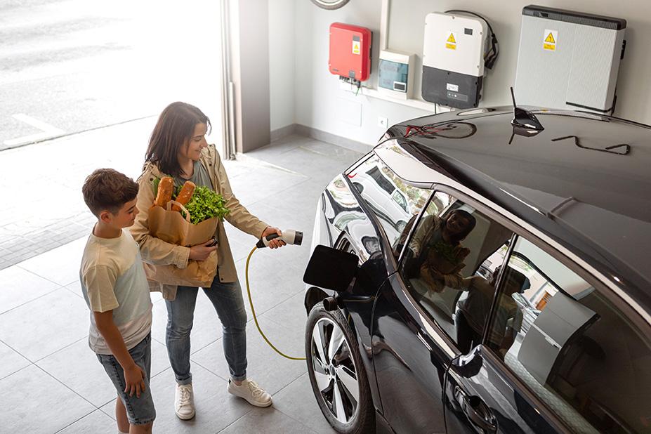 A mom holding groceries charges her electric vehicle with her son standing nearby. They are in a garage.