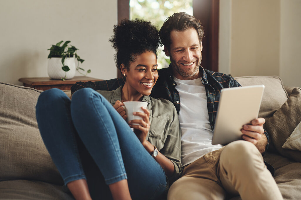 Relax, home and interracial couple on a couch, tablet and happiness with social media and connection Partners, man and woman on a sofa, technology and loving with a smile, romance and online reading